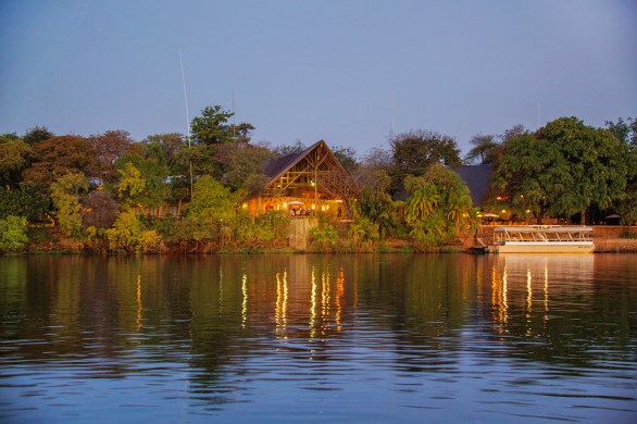Evening view of Chobe Safari Lodge from across the Chobe River, with warm lights reflecting on the water and a moored river safari boat to the right.