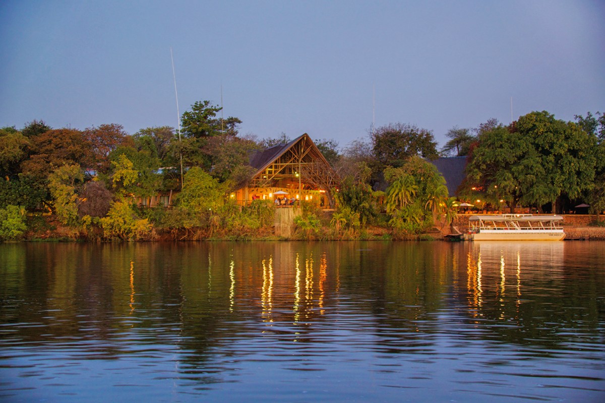 Evening view of Chobe Safari Lodge from across the Chobe River, with warm lights reflecting on the water and a moored river safari boat to the right.